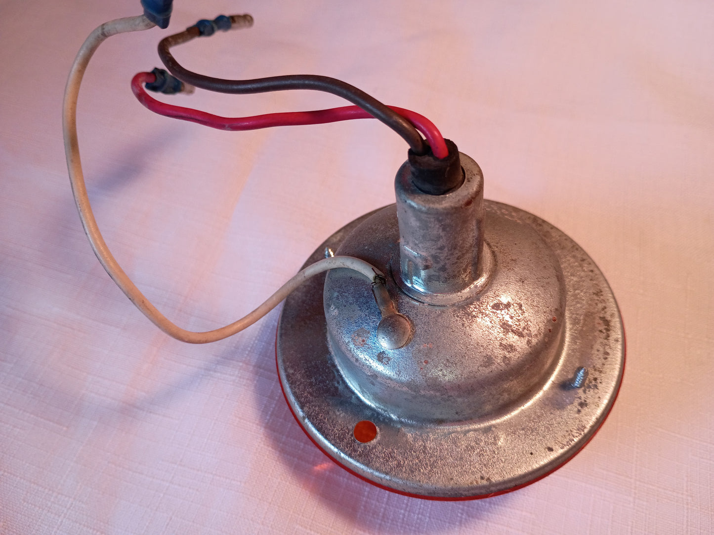 A vintage metal electric lamp base or socket with a circular tarnished silver plate, featuring red and black wires and a beige wire, resting on a light pinkish-beige textured surface.