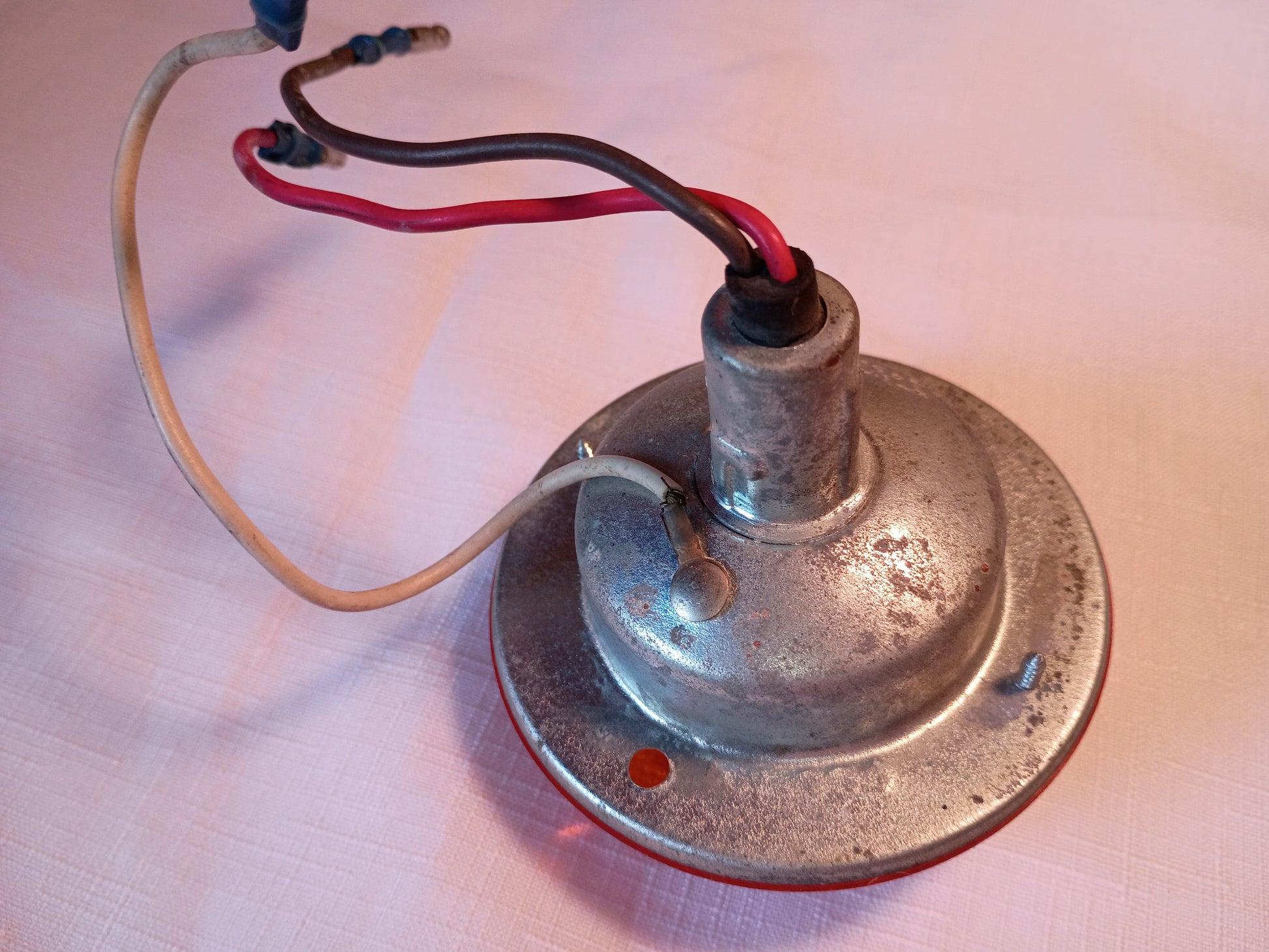 A vintage metal electric lamp base or socket with a circular tarnished silver plate, featuring red and black wires and a beige wire, resting on a light pinkish-beige textured surface.