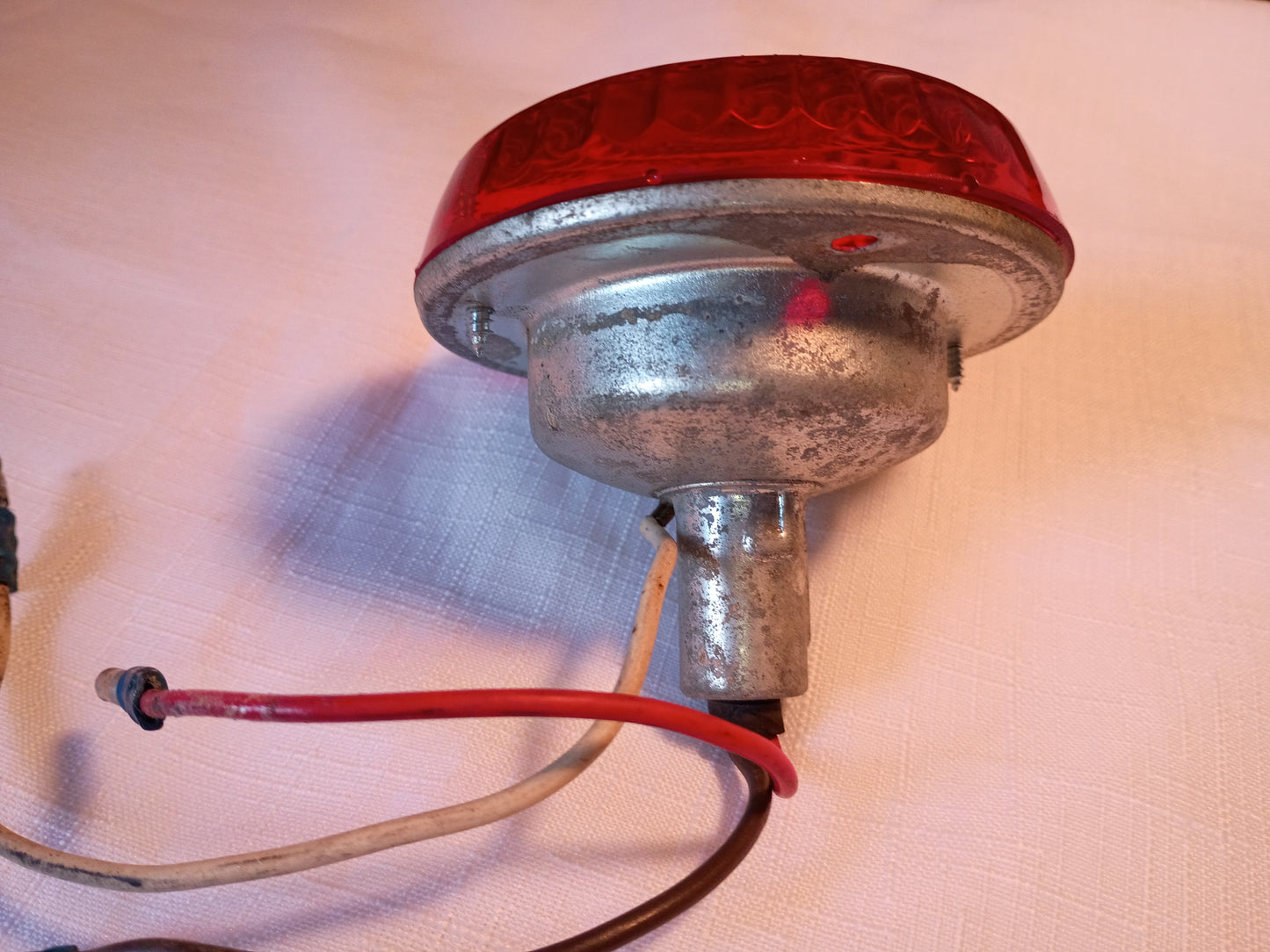 A vintage red rear marker light with a galvanized metal housing and attached red, white, and brown wires, resting on a light-colored textured fabric background.