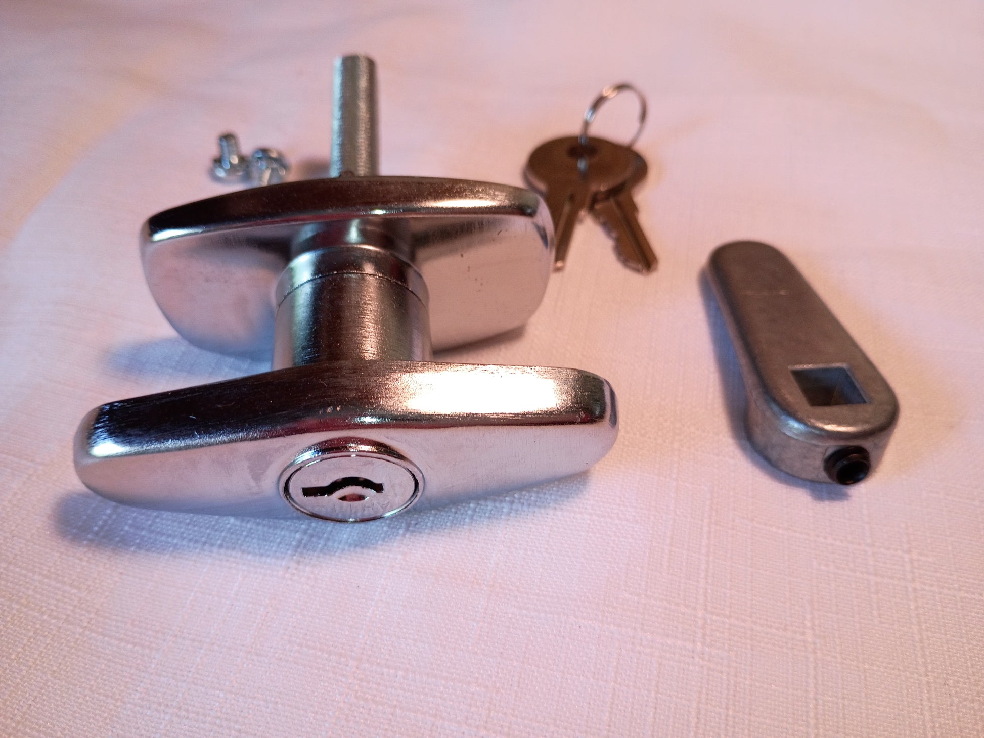 A polished chrome T-bar lock with a matching key, shown alongside a small gray electronic lock pick or lock component on a white textured fabric background.
