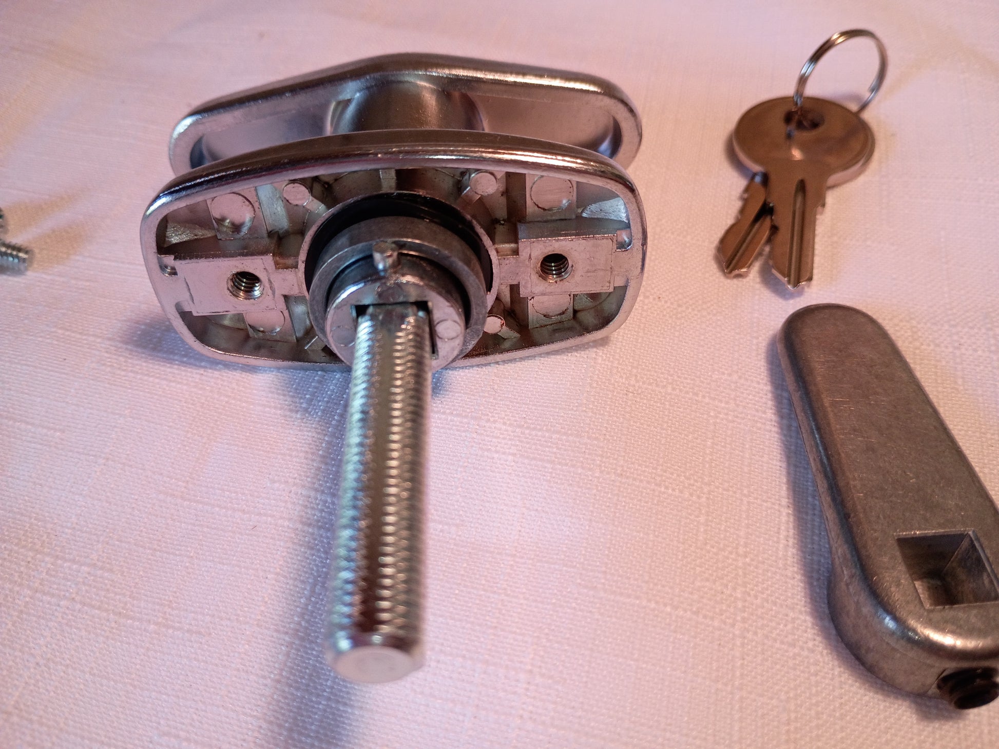 A metal latch or lock hardware component with a long threaded rod, shown with two keys and a matching metal handle piece on a light fabric surface.