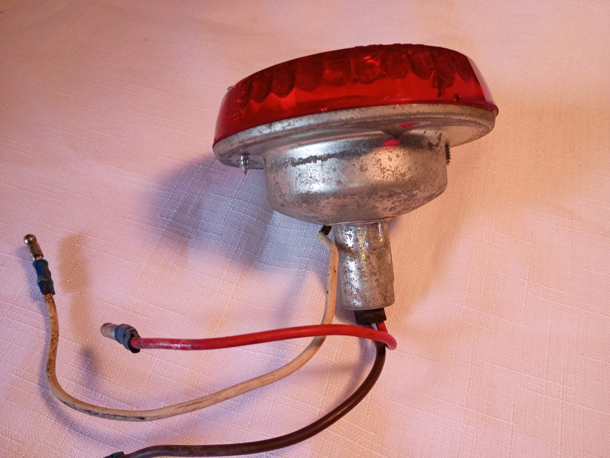A vintage red taillight with a silver metal housing and attached wires, photographed on a light pinkish-beige textured fabric background.