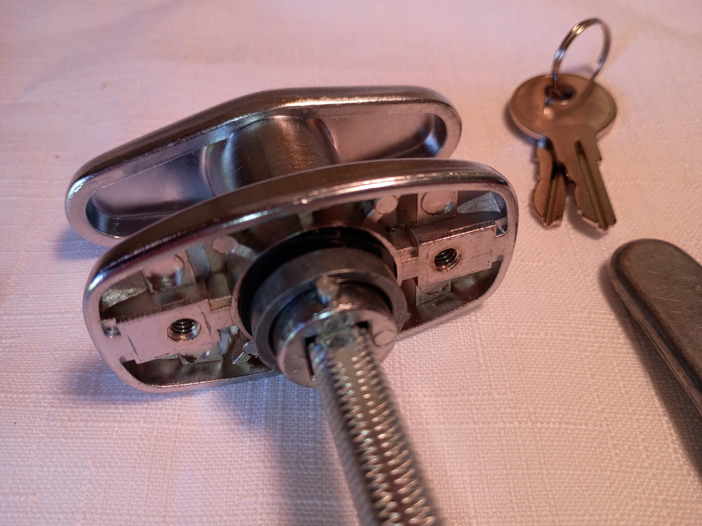 A close-up of a metal latch or lock mechanism with a threaded rod extending from the center, shown with two keys on a white textured surface.