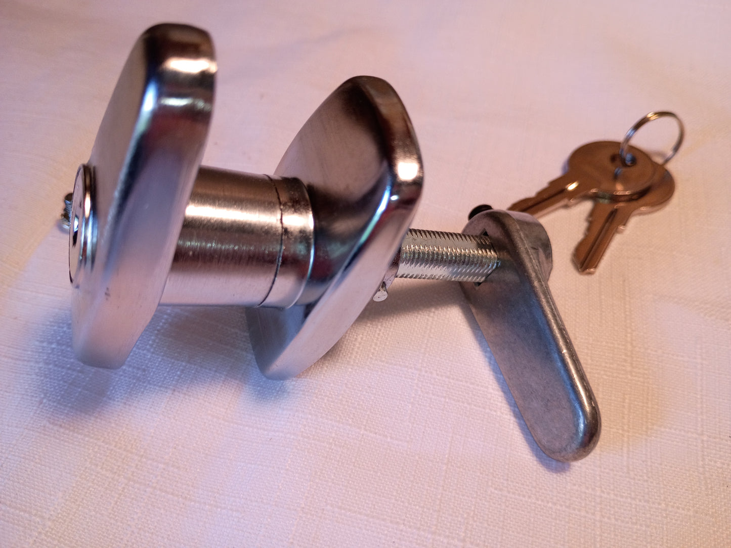 A metallic deadbolt lock with a thumb turn and two keys, displayed on a light-colored textured fabric background.
