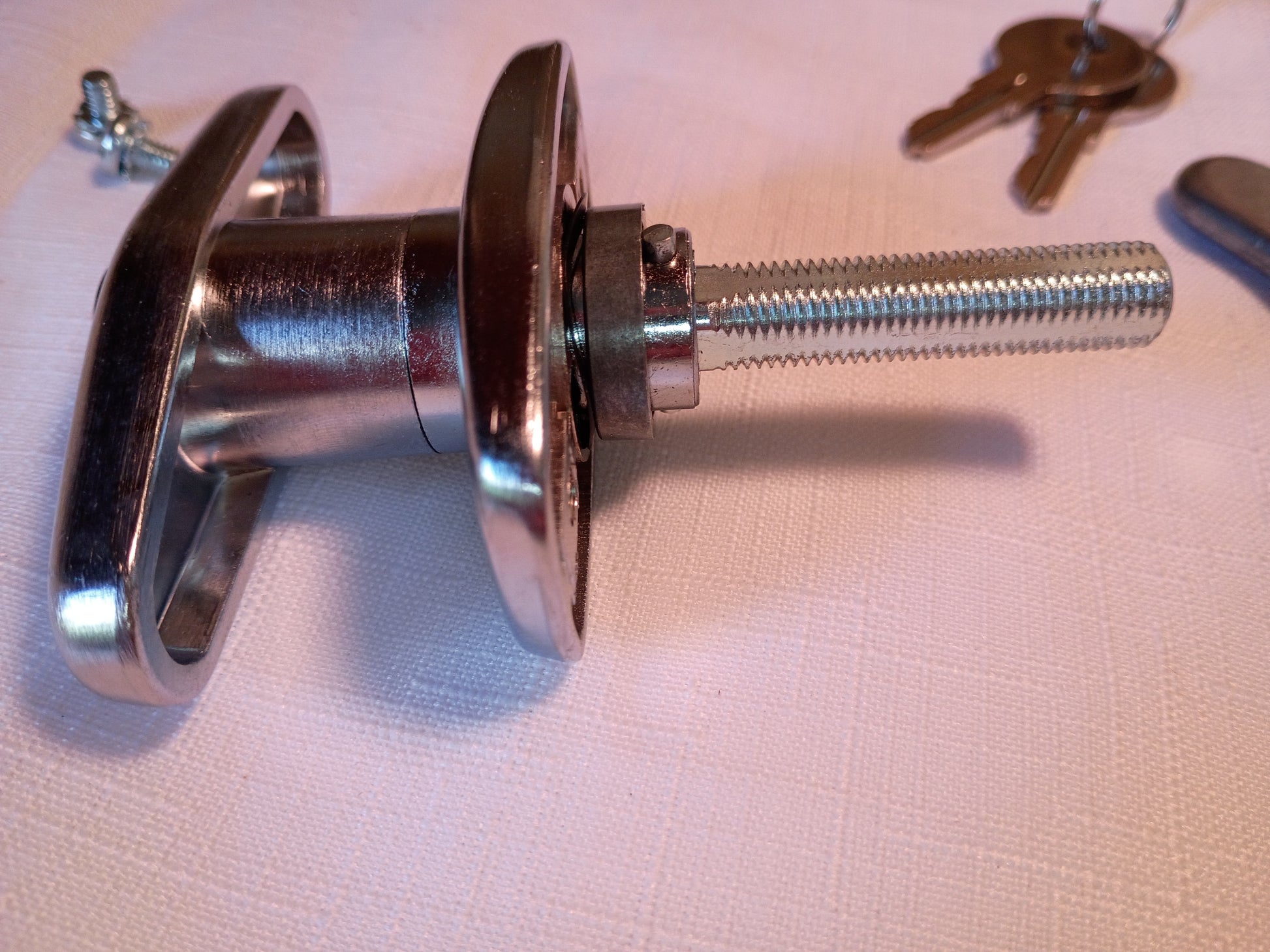 A polished metal door lock cylinder with a threaded bolt shaft and a round thumb turn, shown on a light textured surface with keys in the background.